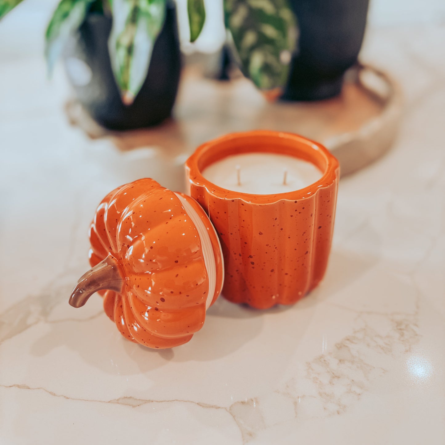 Pumpkin-shaped candle on a marble surface with plants in the background
