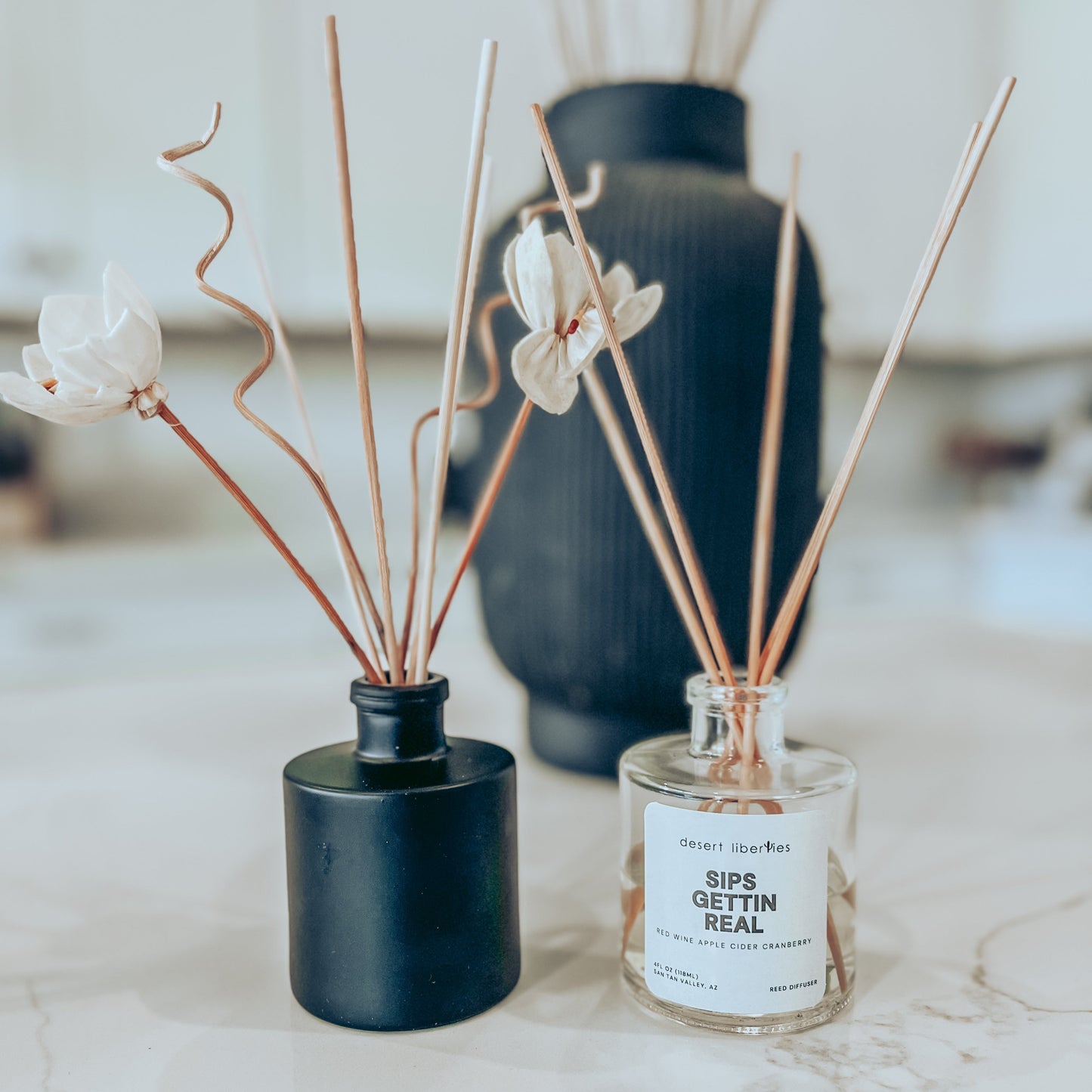 Two reed diffusers. One with dark reeds in a black round jar, and another with light reeds in a clear round jar with a white label.
