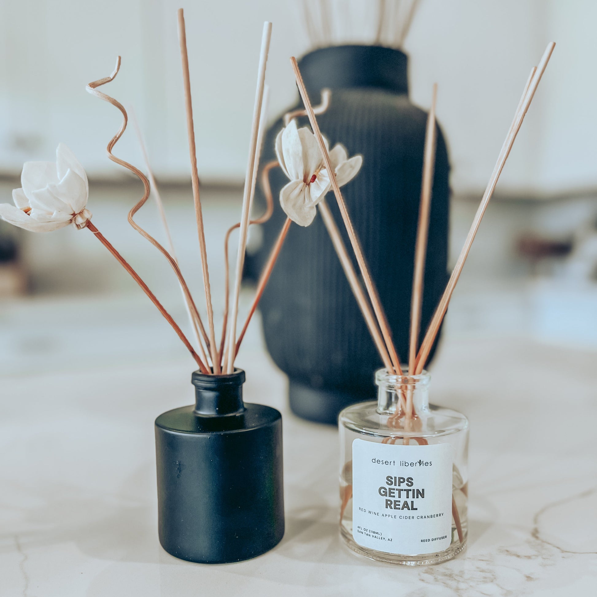 Two reed diffusers. One with dark reeds in a black round jar, and another with light reeds in a clear round jar with a white label.