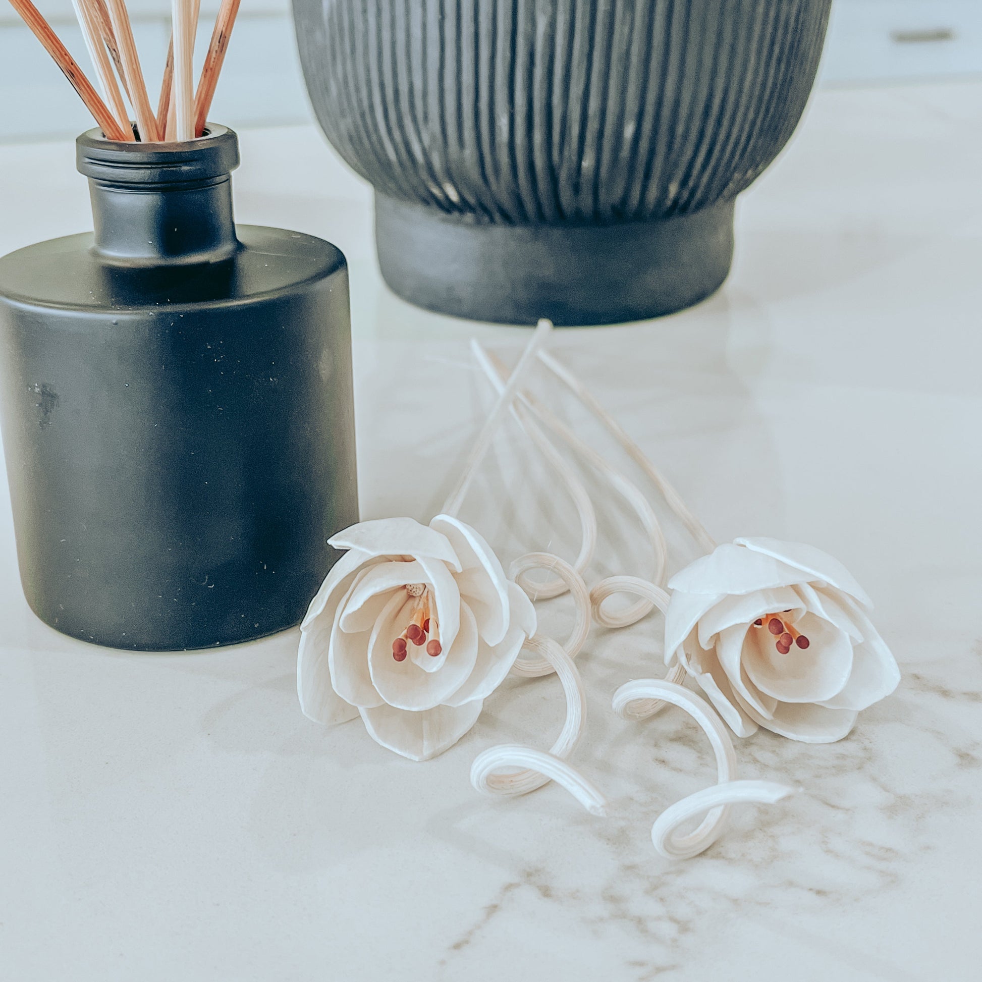 Flower and spiral ornamental reeds on a marble surface with a matte black reed diffuser in the background.
