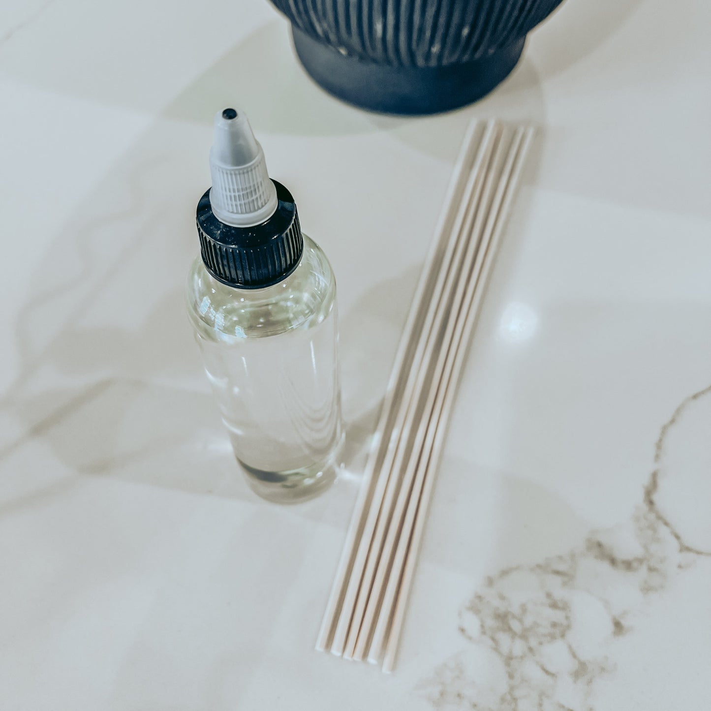 A clear plastic bottle with a black spray nozzle, accompanied by six light brown rattan reeds placed on a white surface.