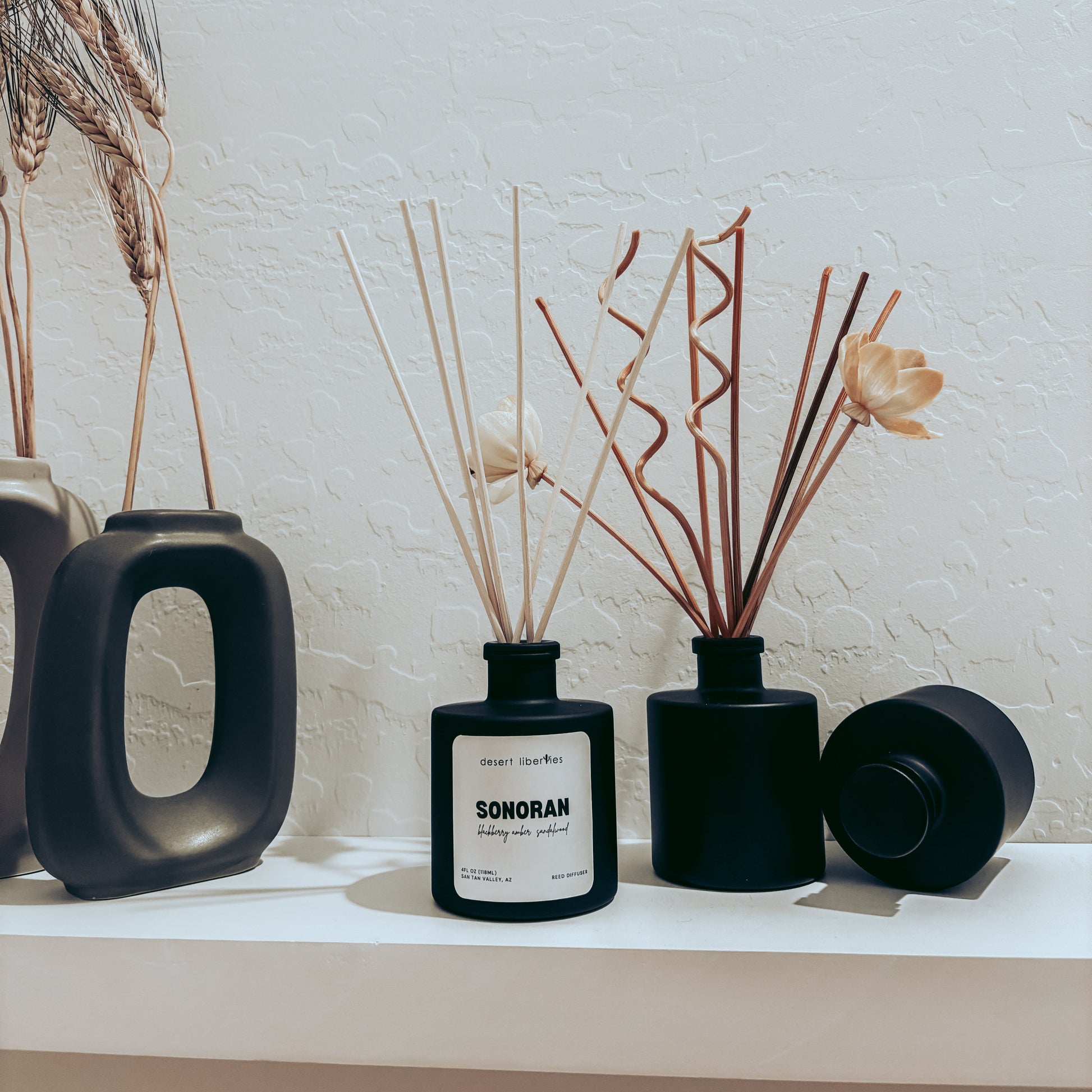 Black vases with dried plants on a white surface against a light brick wall.