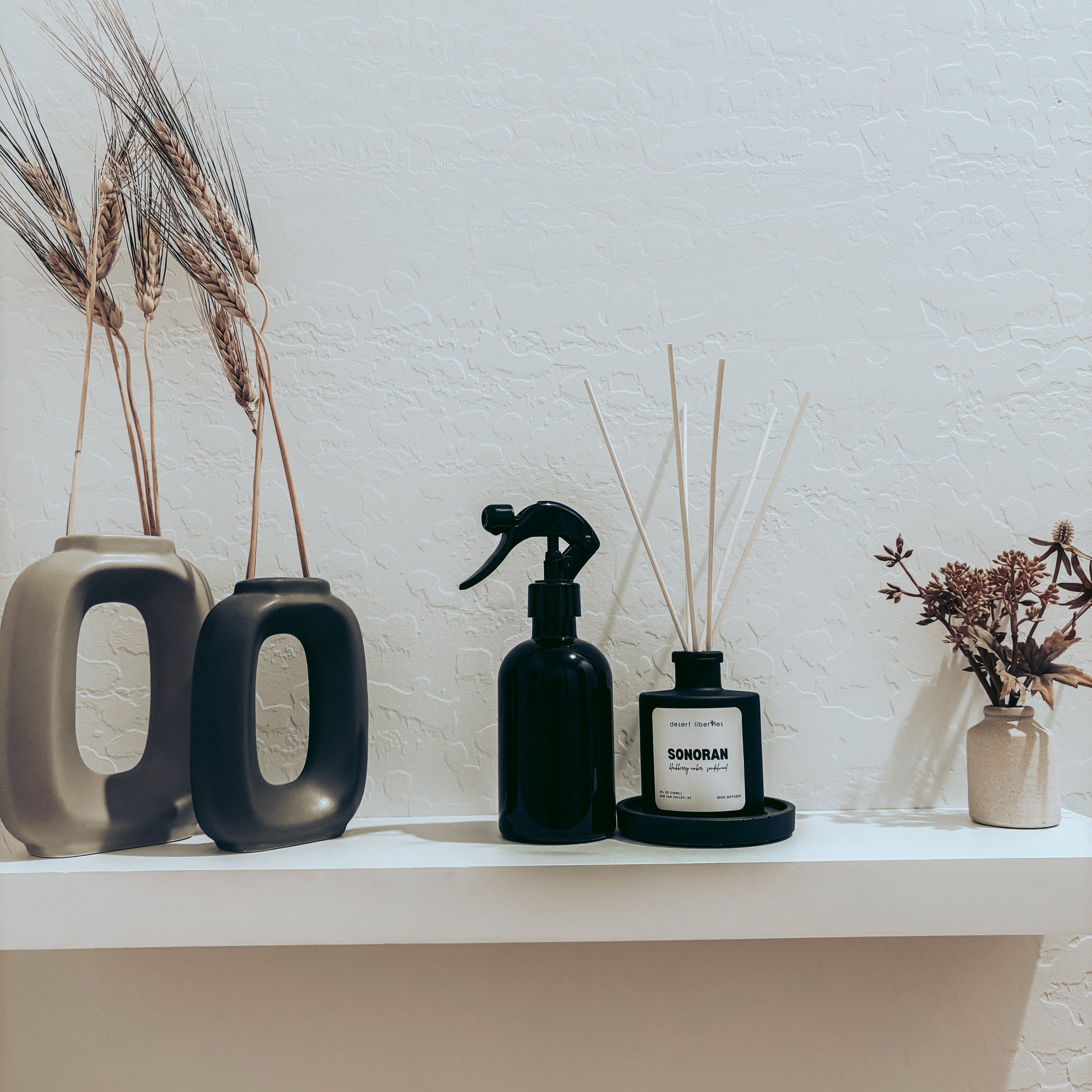 Black spray bottle and diffuser on a white shelf with decorative vases and plants against a light wall.