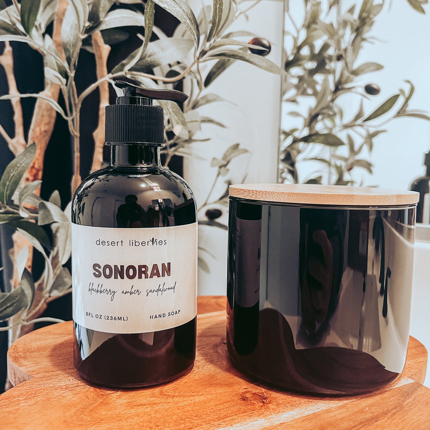 Soap bottle next to an unlabeled candle on a wood surface with olive leaves in the background. 