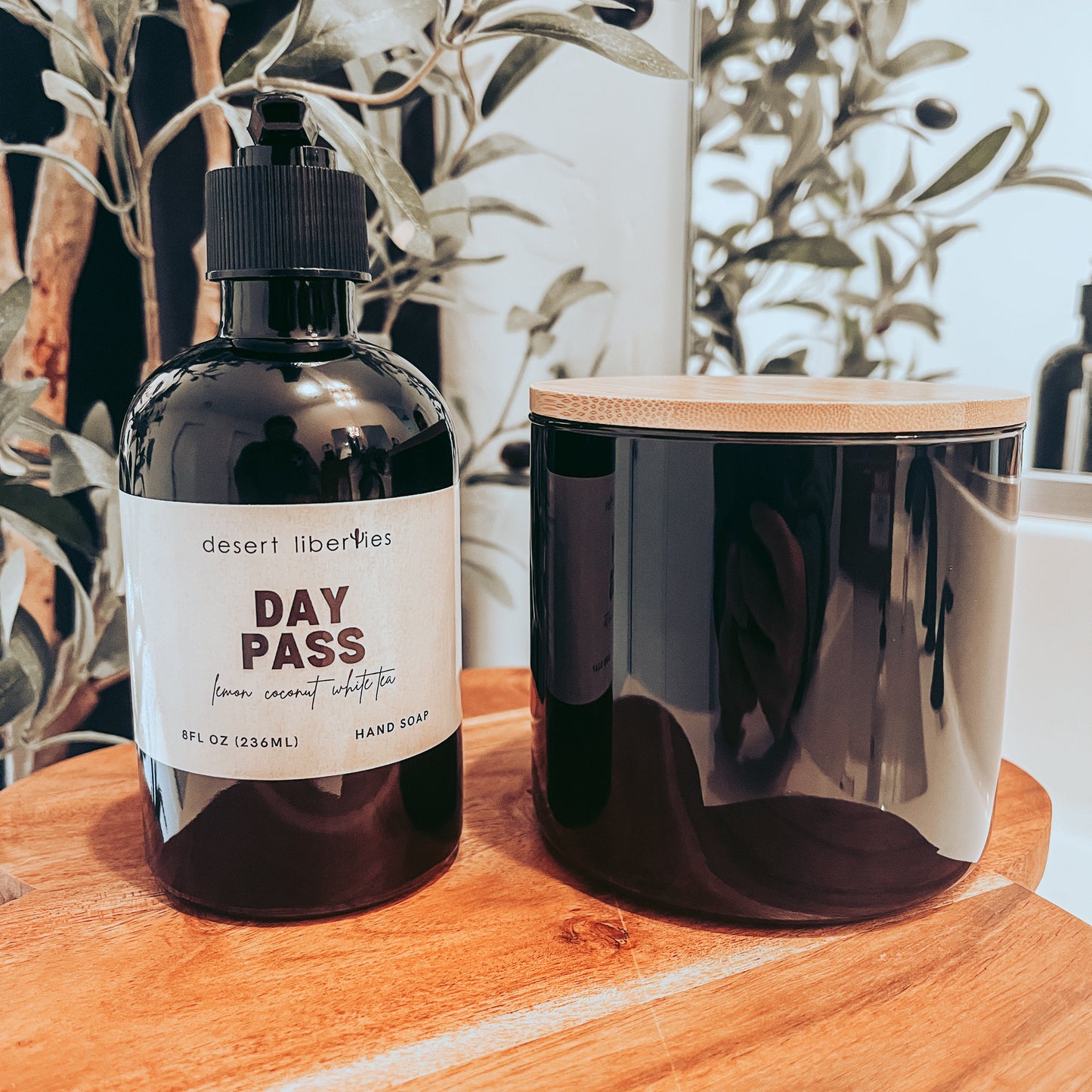 Soap bottle next to an unlabeled candle on a wood surface with olive leaves in the background. 