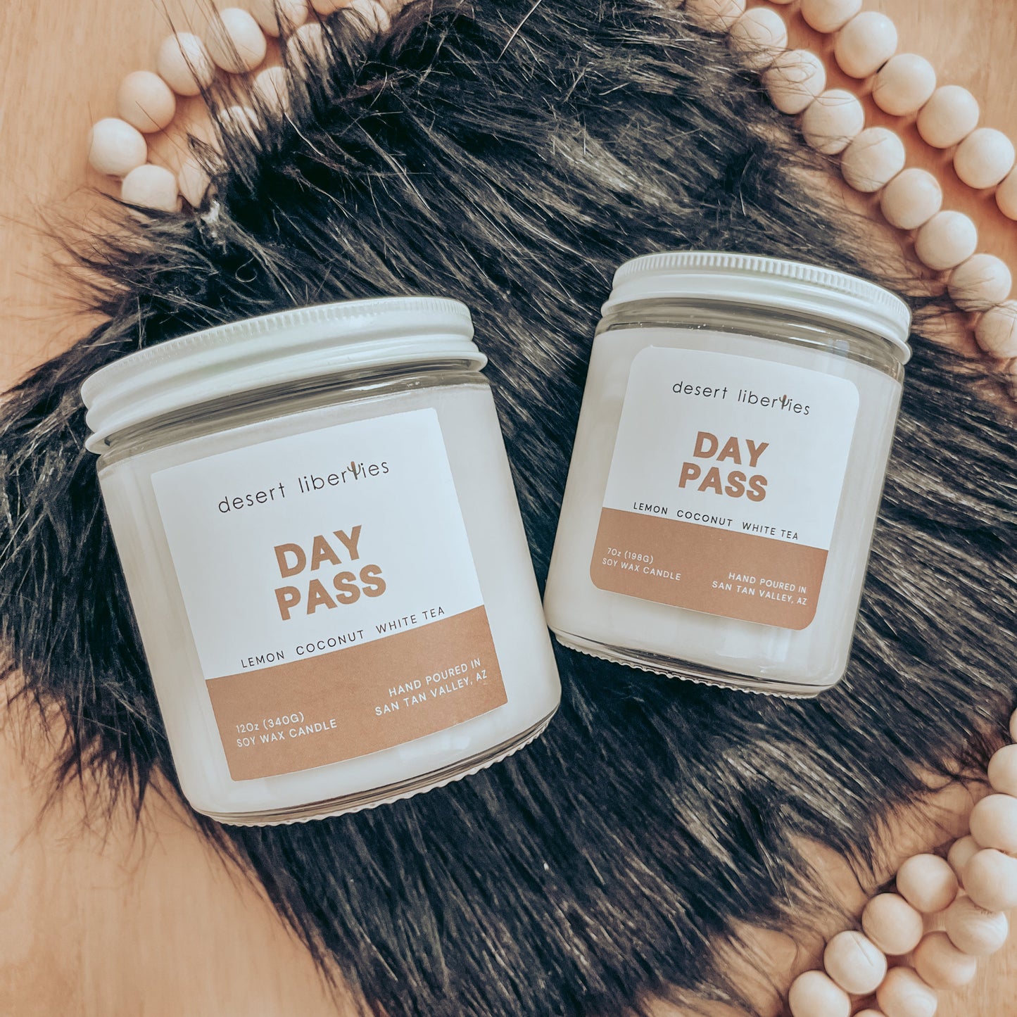 Two candles with white labels, one larger and one smaller, both labeled 'DAY PASS' with fragrance notes listed below the name. The candles are placed on a surface with a fur-like texture and wooden beads.