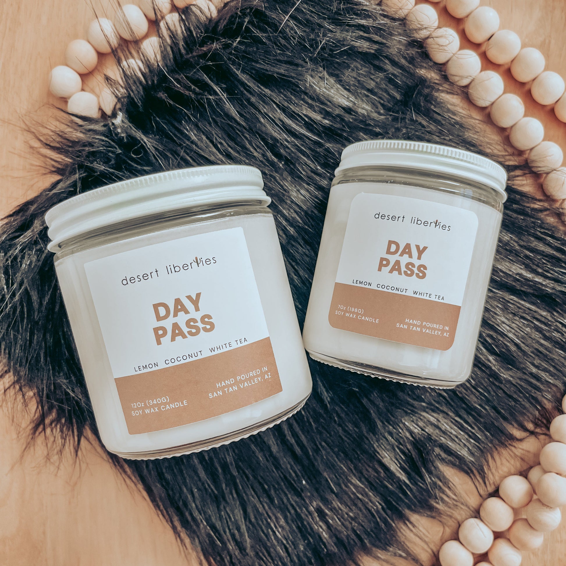 Two candles with white labels, one larger and one smaller, both labeled 'DAY PASS' with fragrance notes listed below the name. The candles are placed on a surface with a fur-like texture and wooden beads.