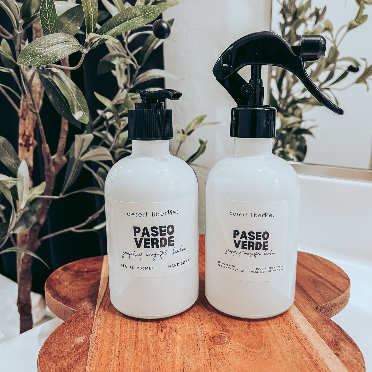 Soap bottle next to a matching spray bottle on a wood surface and olive tree in the background. 