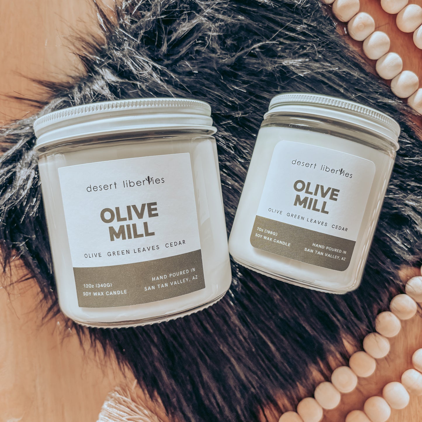 Two candles labeled 'Olive Mill' by Desert Liberties on a textured surface with beads.