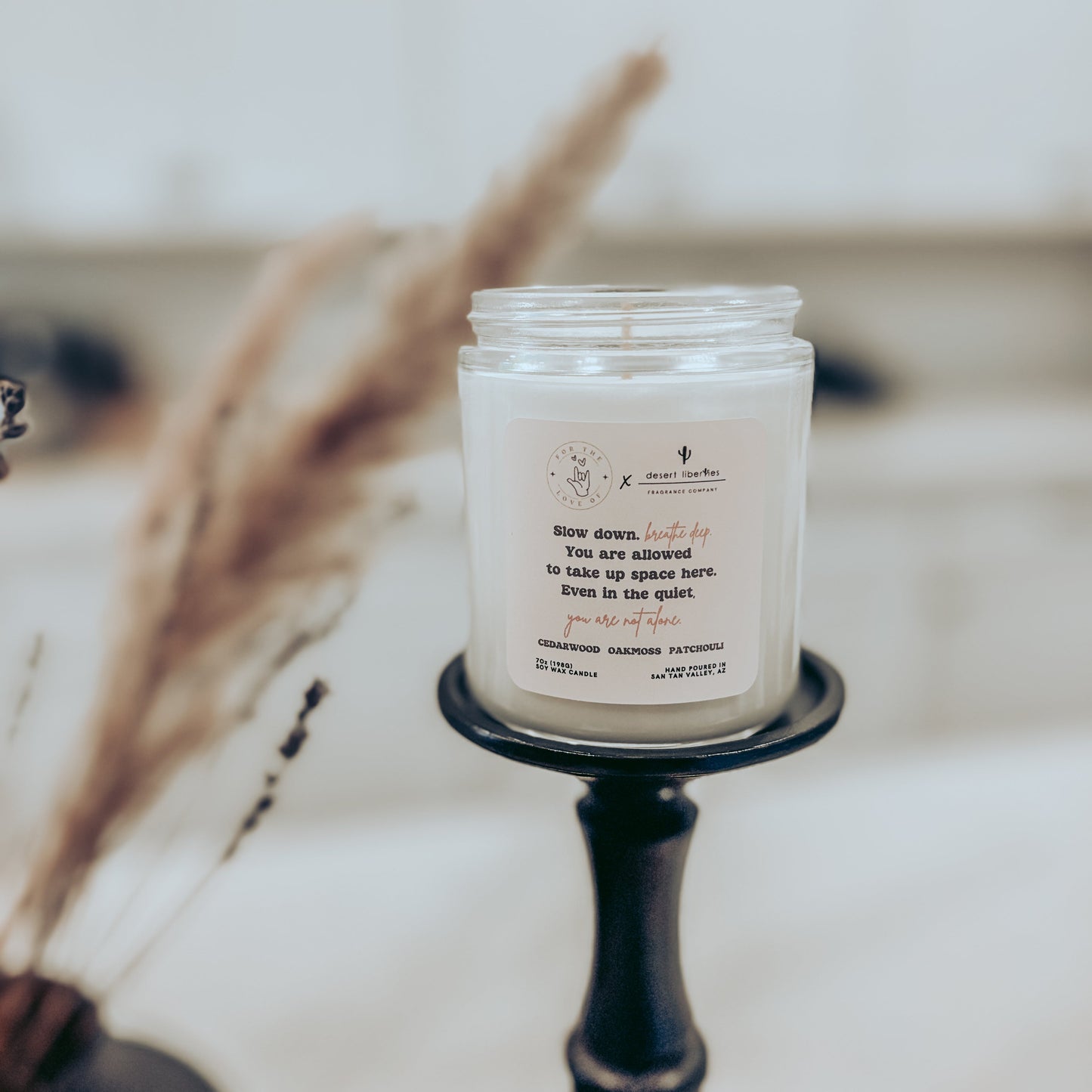 Candle in a glass jar with a label on a stand, surrounded by dried plants.