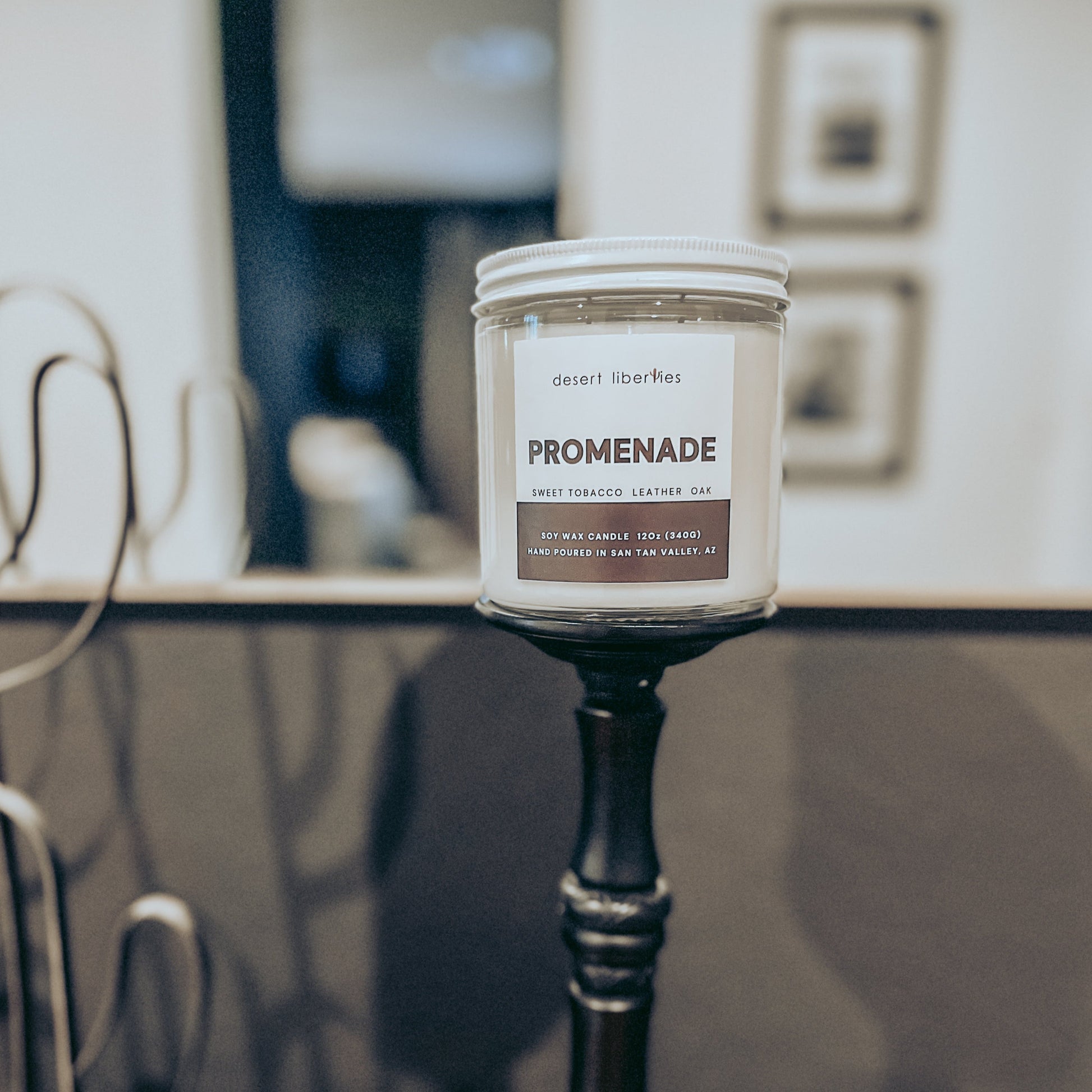 A candle with a white label labeled 'PROMENADE' on a vessel, set against a blurred background.