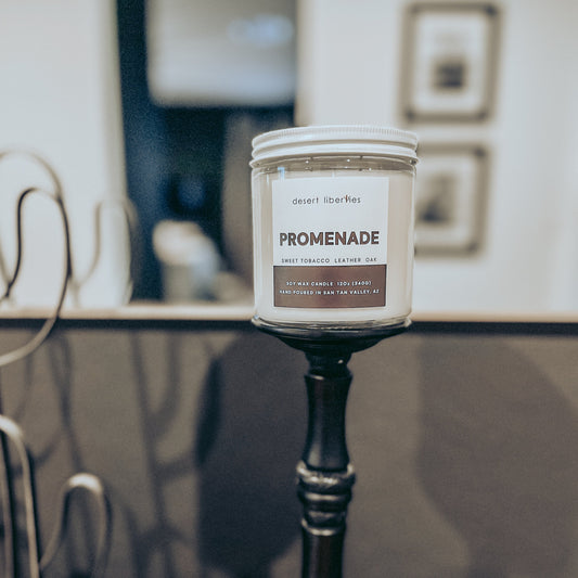 A candle with a white label labeled 'PROMENADE' on a vessel, set against a blurred background.