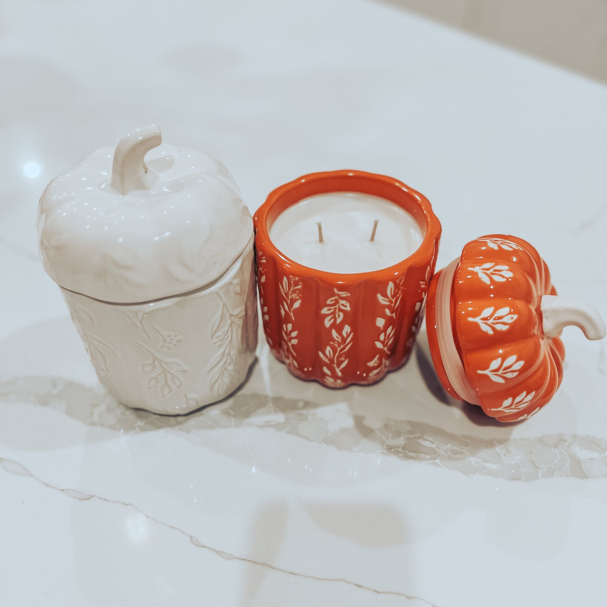 A white candle in a ceramic pumpkin container with a pumpkin design, and a red candle in a ceramic pumpkin container with a leaf pattern. Both are on a marble surface.