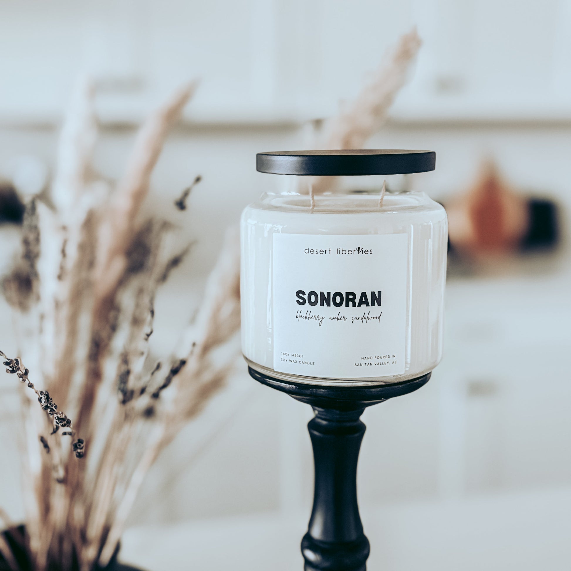 A white soy candle in a clear glass jar with a black lid, labeled 'SONORAN' by Desert Liberties, displayed on a black candlestick holder in a softly blurred indoor setting with dried grasses in the foreground.