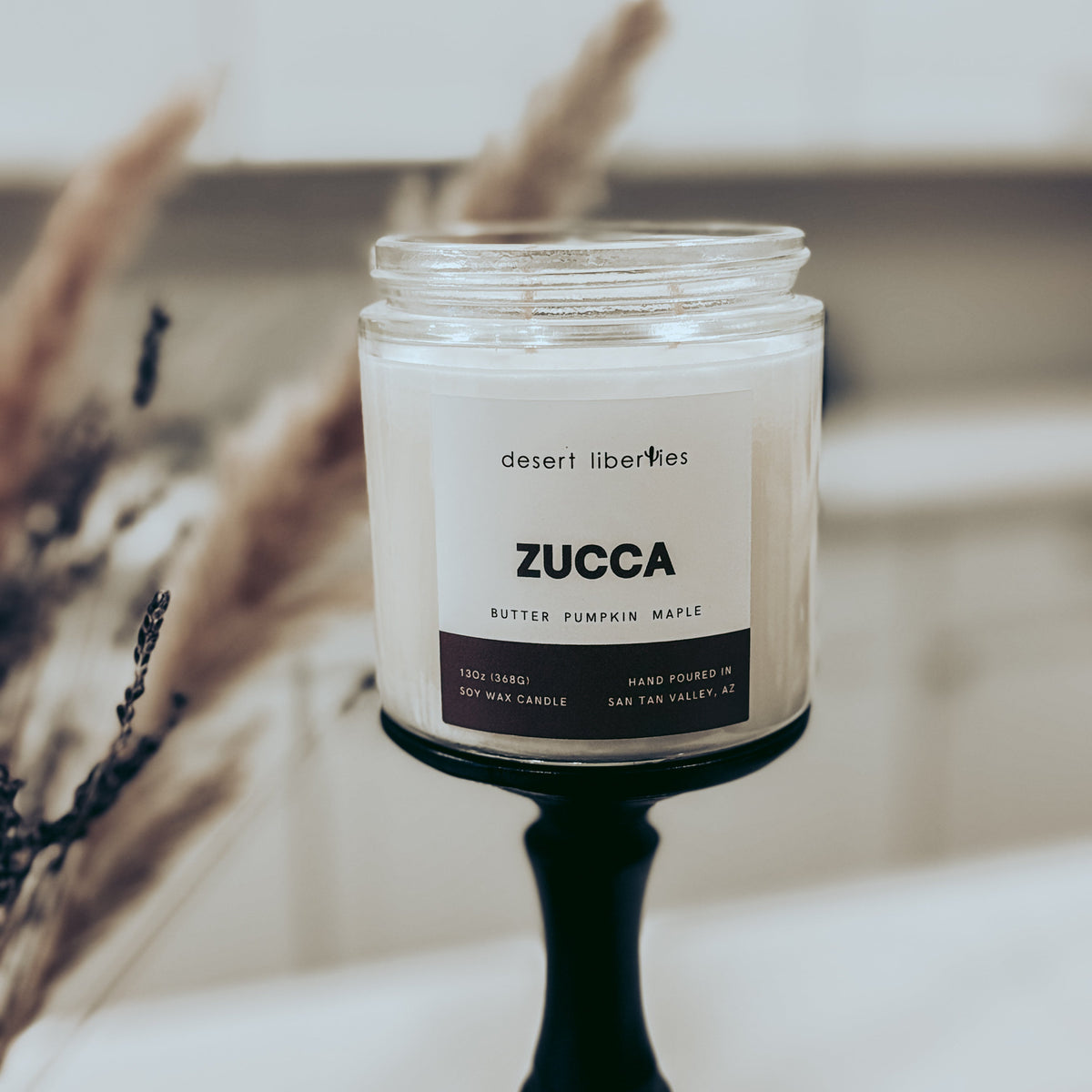 A jar candle labeled 'ZUCCA' by desert liberties, set on a black pedestal with blurred dried grass in the background.