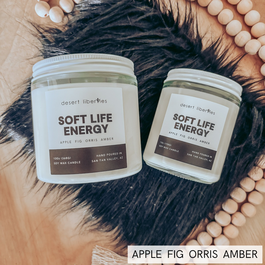 Two glass jar candles labeled 'desert liberies SOFT LIFE ENERGY' are arranged on a wood surface with a black feather and a circular arrangement of light wood beads. A text overlay at the bottom reads 'APPLE FIG ORRIS AMBER'.