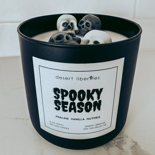 A black candle jar labeled 'desert liberties Spooky Season' with three skull-shaped wax ornaments floating on the cream-colored wax inside, set on a light marble surface against a white tiled background.