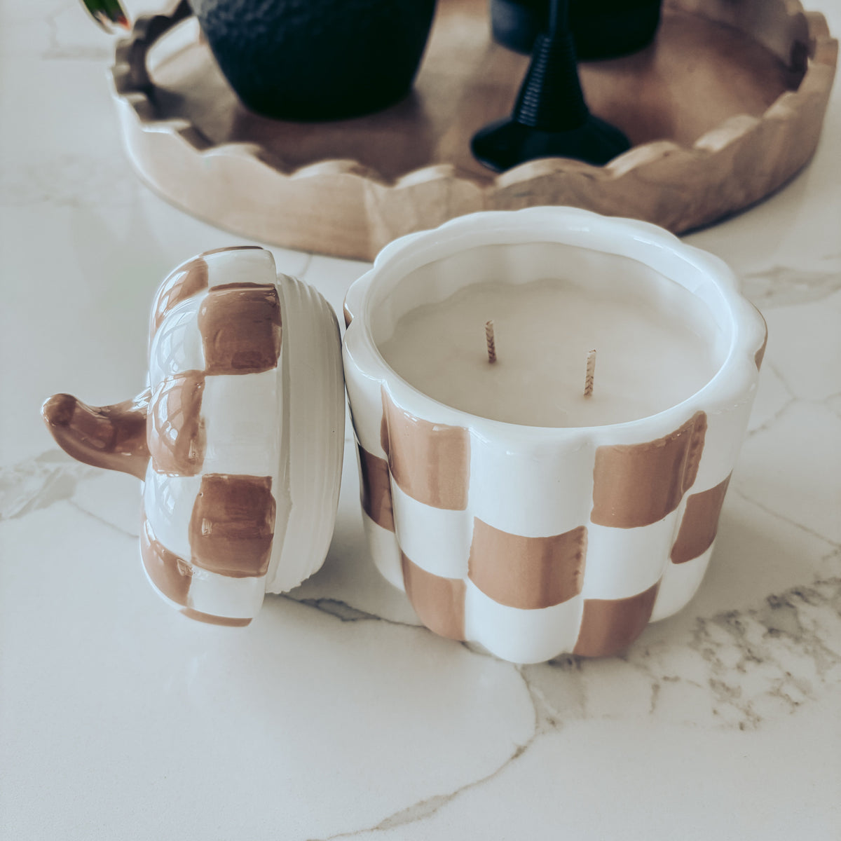 A ceramic candle jar with a lid, featuring a brown and white checkered pumpkin design, set on a white marble countertop with a wooden tray and potted plants in the background.