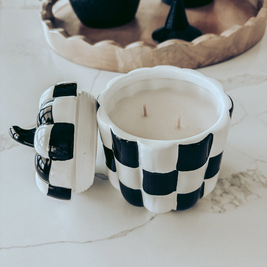 A black and white checkered ceramic candle jar with its lid off, showing two wicks, set on a light marble surface with a decorative tray and black vases in the background.