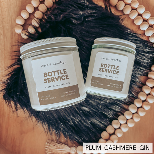 Two glass jar candles labeled 'Bottle Service' by desert liberties, styled on a wooden tray with a black faux-fur pad and a circular wooden bead border. Text overlay reads 'PLUM CASHMERE GIN'.