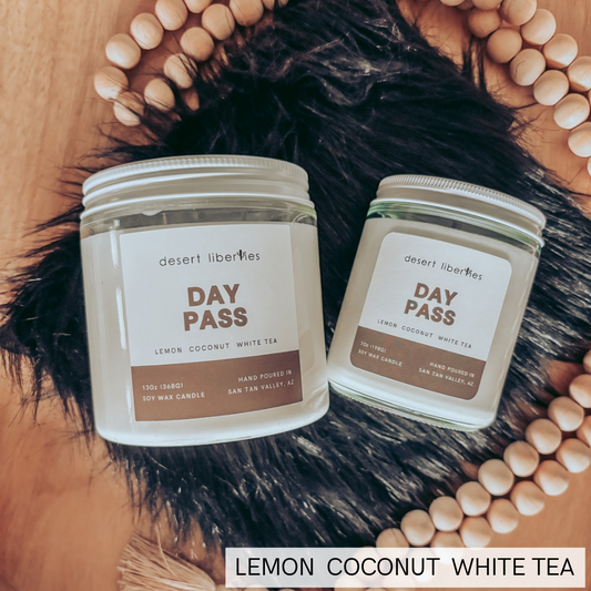 Two glass jar candles labeled 'desert liberties DAY PASS' and 'LEMON COCONUT WHITE TEA' arranged on a black faux-fur surface with a wooden tray, wooden beads, and tassels.