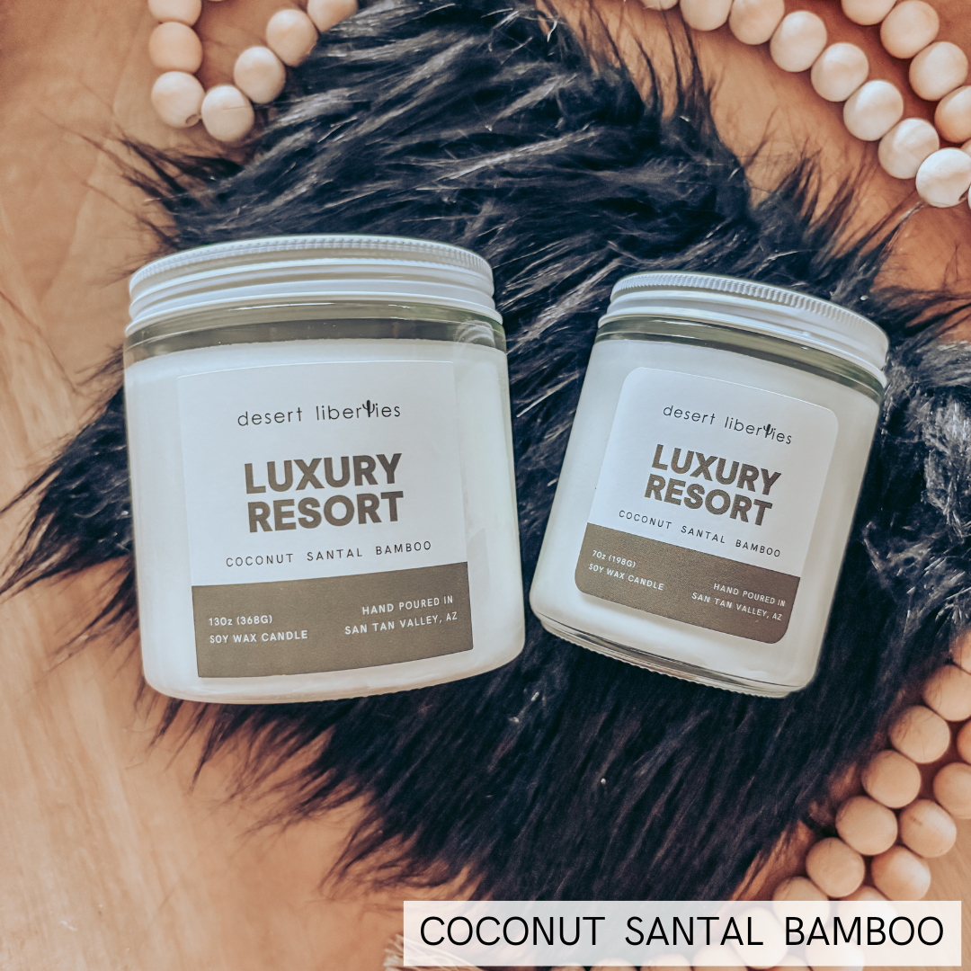 Two glass jars of Desert Liberties 'Luxury Resort' coconut santal bamboo scented candles are arranged on a black faux fur rug over a wooden tray, surrounded by a circular beaded necklace.
