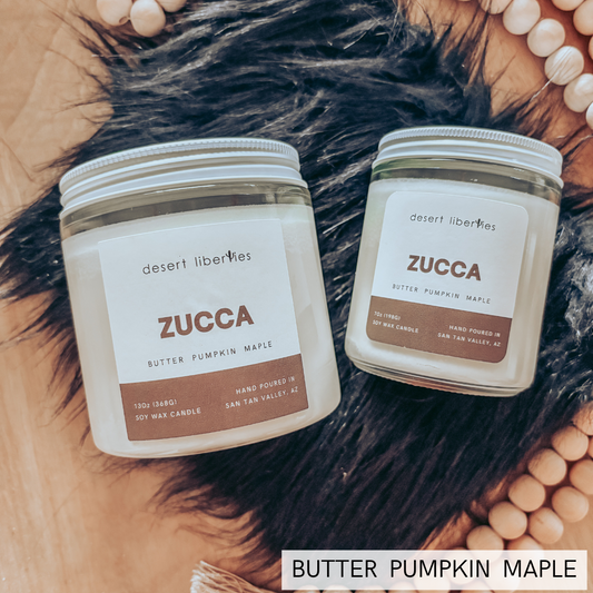 Two glass jar candles on a wooden surface with a black faux-fur prop and peach-colored beads. A text overlay at the bottom reads 'BUTTER PUMPKIN MAPLE'.