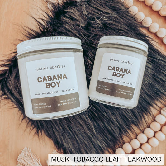 Two 'Cabana Boy' candles on a textured surface with beads and fur.