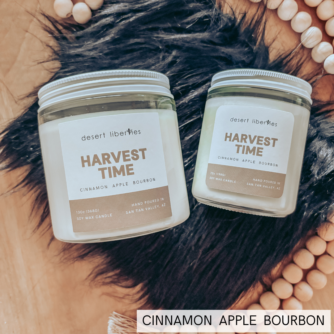 Two Desert Liberties 'Harvest Time' scented candles in glass jars w, displayed on a wooden surface with black faux fur, beads, and a white text banner reading 'CINNAMON APPLE BOURBON'.
