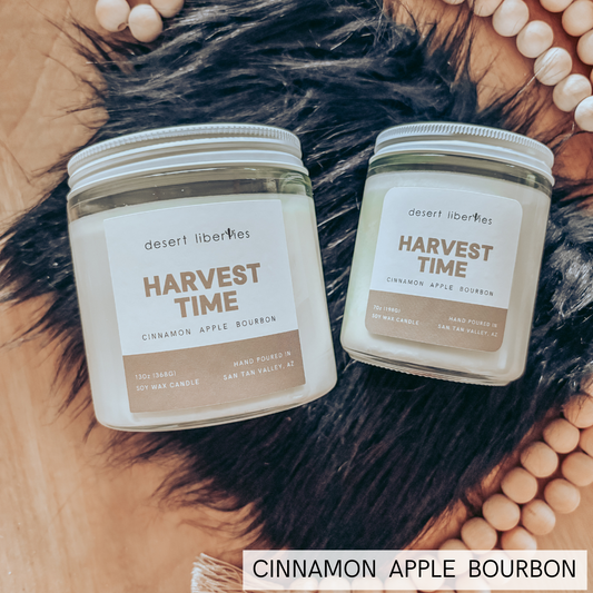 Two Desert Liberties 'Harvest Time' scented candles in glass jars w, displayed on a wooden surface with black faux fur, beads, and a white text banner reading 'CINNAMON APPLE BOURBON'.