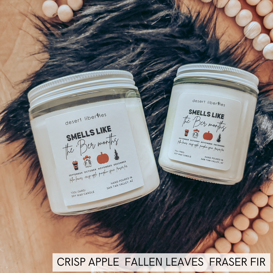 Two white scented candles in clear glass jars with white lids, arranged on a black faux-fur rug with wooden beads on a tan surface. Text at the bottom reads 'CRISP APPLE FALLEN LEAVES FRASER FIR'.