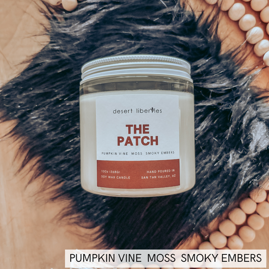 A jar candle labeled 'THE PATCH' by desert liberties, placed on a black faux-fur surface with a tan woven mat and wooden beads around it. The label reads 'Pumpkin Vine Moss Smoky Embers.