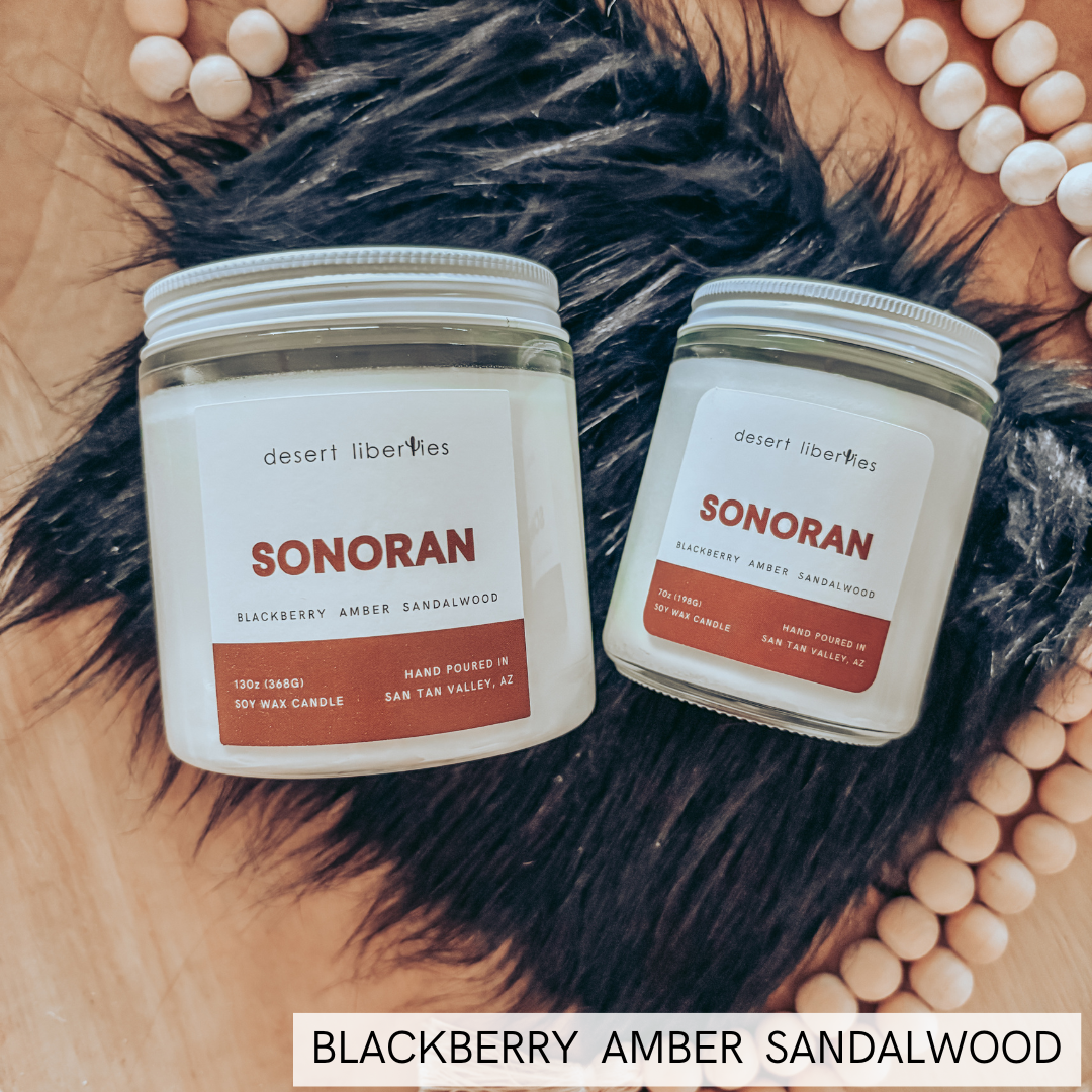Two candles in glass jars arranged on a wooden surface with black faux fur and a beaded necklace. A text overlay at the bottom reads 'BLACKBERRY AMBER SANDALWOOD'.